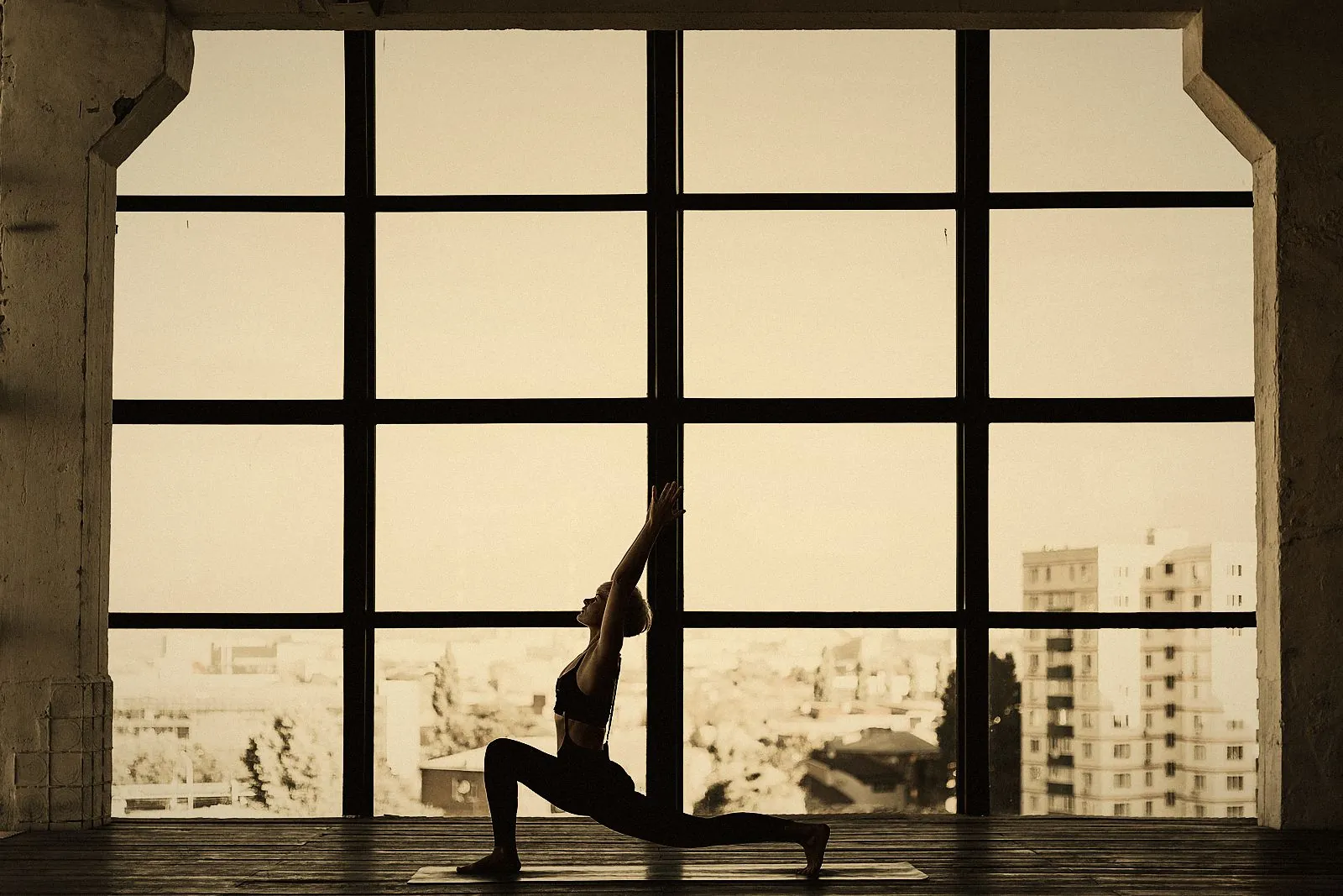 A low lunge yoga pose in front of tall windows overlooking the city